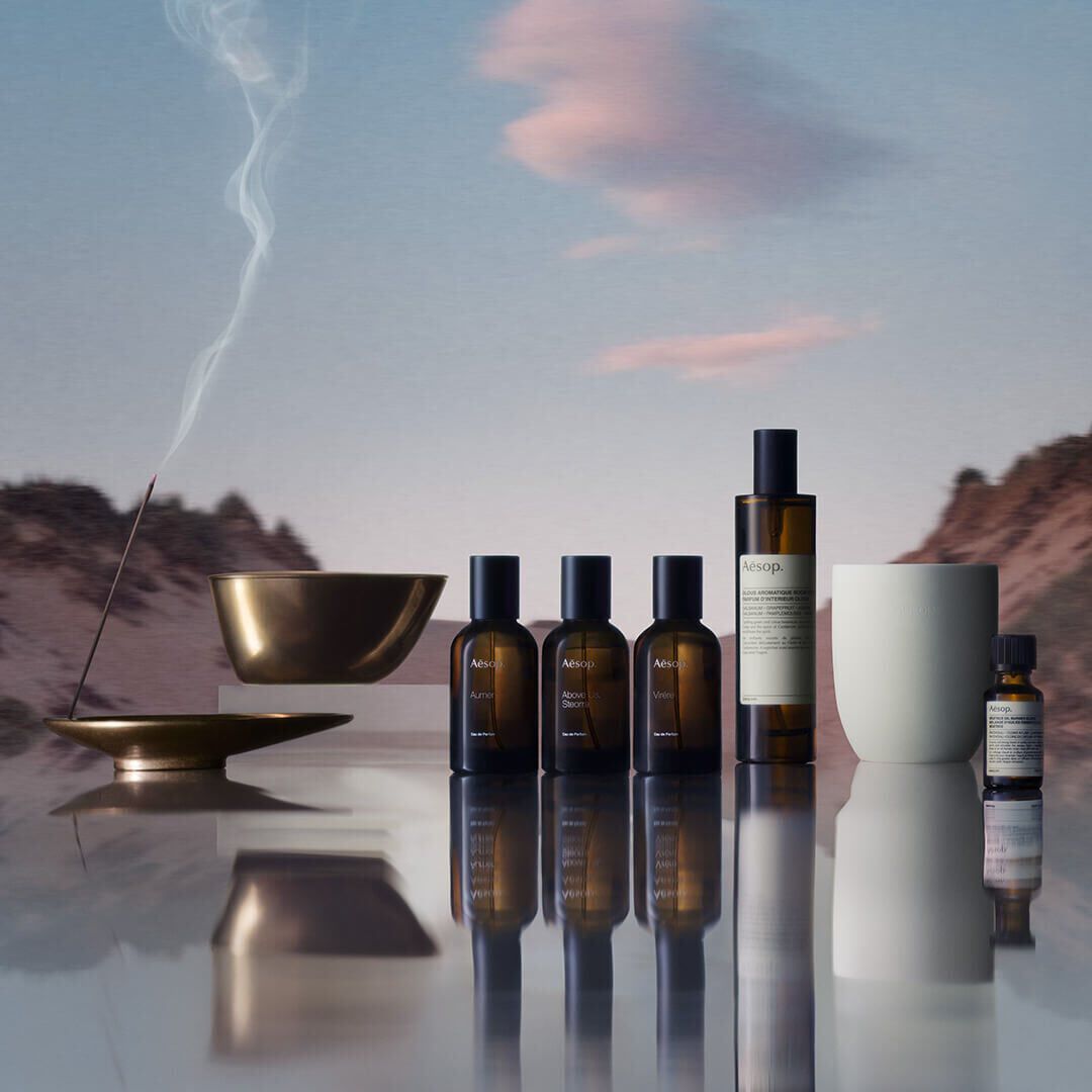 Eine Kollektion von Aesop-Formulierungen, darunter Eaux de Parfum, Raumsprays und atherische Ole, ein Raucherstabchen, das in einem bronzenen Halter brennt, vor einer verschwommenen Berglandschaft, die sich auf einer Oberflache spiegelt.