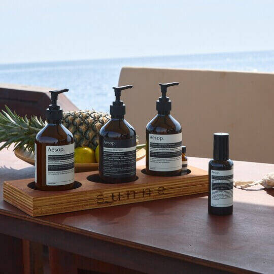 Five Aesop formulations and tropical fruit on a table by the beach.
